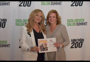Two women stand in front of a "Data-Driven Salon Summit" backdrop, smiling at the camera while holding a certificate and a Salon Today 200 magazine. - Mermaid Hair Extensions in Kirkland and Seattle, WA