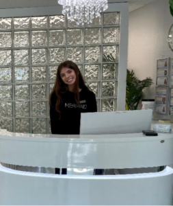 A woman stands behind a white reception desk, smiling, with a glass block wall and a hanging light fixture in the background at BeamBell Salon, known for its expert Mermaid Hair Extensions. - Mermaid Hair Extensions in Kirkland and Seattle, WA